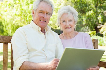 Man and a woman looking ahead as they hold a laptop between them while sitting on a park bench Stock Photo - Premium Royalty-Free, Code: 6109-06004633
