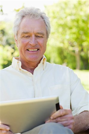 Homme souriant tout en regardant l'écran d'une tablette, assis sur un banc dans le parc Photographie de stock - Premium Libres de Droits, Code: 6109-06004734