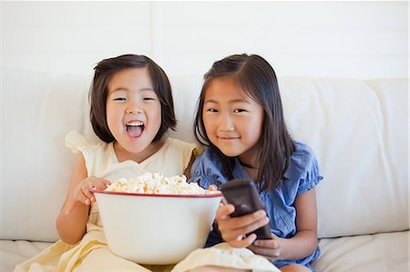 smiling kid eating bowl - Two laughing sisters watching tv together with a bowl of popcorn and the tv remote Stock Photo - Premium Royalty-Free, Code: 6109-06004845