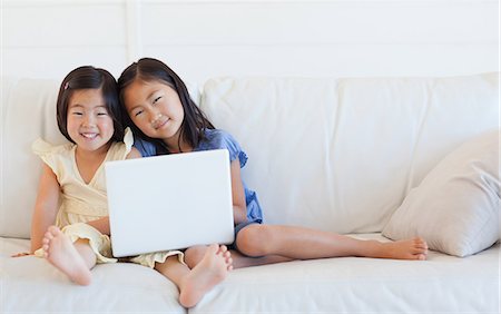 Two sisters looking forward and smiling while holding a laptop on the couch Stock Photo - Premium Royalty-Free, Code: 6109-06004835