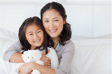 pretty mother - A daughter with a teddy in her hands smiles as her mother holds her close while they sit on the bed Stock Photo - Premium Royalty-Free, Code: 6109-06005021