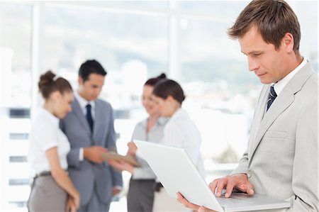 simsearch:6109-06005688,k - Young salesman working his laptop with colleagues behind him Foto de stock - Royalty Free Premium, Número: 6109-06005684