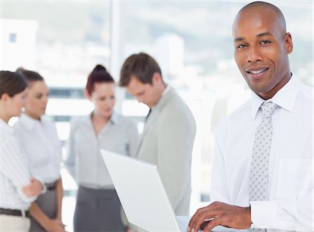 simsearch:6109-06005688,k - Smiling young salesman with his laptop and associates behind him Foto de stock - Royalty Free Premium, Número: 6109-06005741