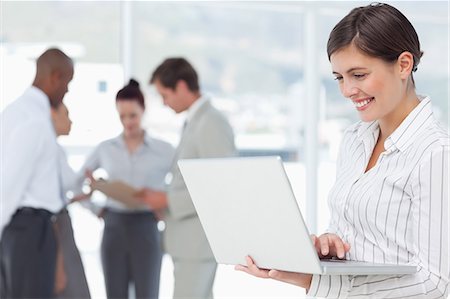 simsearch:6109-06005753,k - Smiling young saleswoman with laptop and colleagues behind her Stock Photo - Premium Royalty-Free, Code: 6109-06005718
