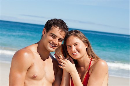Portrait d'un couple souriant écoutant un coquillage avec la mer en arrière-plan Photographie de stock - Premium Libres de Droits, Code: 6109-06005992
