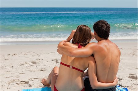 embrace couple swim suit - Rear view of a young couple sitting on beach towel while pointing something in front of the sea Stock Photo - Premium Royalty-Free, Code: 6109-06006039