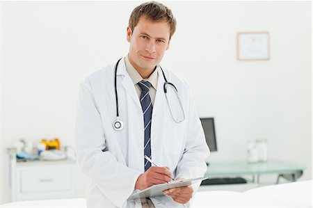 simsearch:6109-06005901,k - Smiling male doctor with clipboard standing in examination room Fotografie stock - Premium Royalty-Free, Codice: 6109-06006355