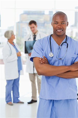 simsearch:6109-06006507,k - Smiling doctor in scrubs with folded arms and two colleagues behind him Fotografie stock - Premium Royalty-Free, Codice: 6109-06006517