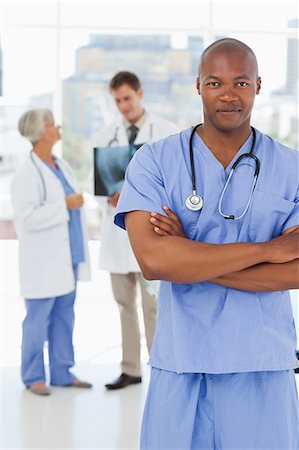 simsearch:6109-06006507,k - Doctor in scrubs with folded arms and two colleagues behind him Fotografie stock - Premium Royalty-Free, Codice: 6109-06006516