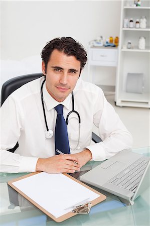 Young doctor sitting at his desk with clipboard and laptop Stock Photo - Premium Royalty-Free, Code: 6109-06006747