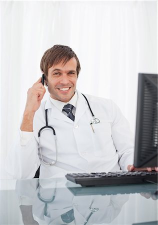 Doctor talking on his mobile phone while smiling and sitting at his desk Stock Photo - Premium Royalty-Free, Code: 6109-06006843