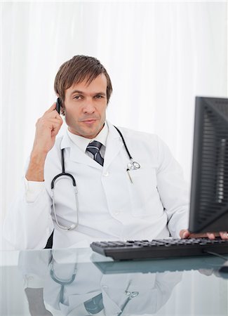 Thoughtful doctor using his mobile phone while raising an eyebrow and sitting at his desk Stock Photo - Premium Royalty-Free, Code: 6109-06006844