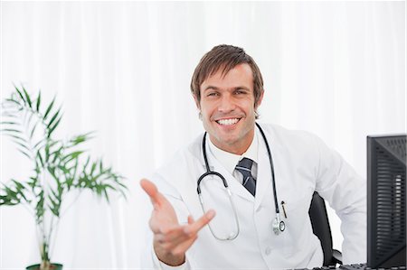 raising her hand - Smiling doctor extending his hand while sitting behind his desk with a computer on it Stock Photo - Premium Royalty-Free, Code: 6109-06006851