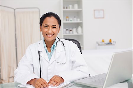 simsearch:6109-06006905,k - Smiling doctor sitting at the desk with a clipboard and a laptop in a medical office Stock Photo - Premium Royalty-Free, Code: 6109-06006897