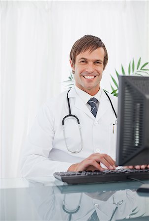 simsearch:6109-06006905,k - Smiling doctor typing on his computer while sitting at a desk in a bright room Stock Photo - Premium Royalty-Free, Code: 6109-06006820