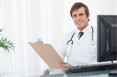 simsearch:6109-06006829,k - Happy doctor looking at the camera while sitting at the desk and holding a clipboard Foto de stock - Sin royalties Premium, Código: 6109-06006833