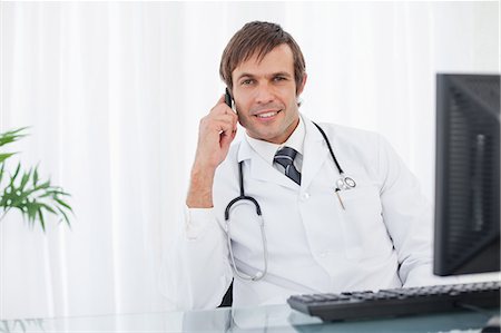 Smiling surgeon sitting at the desk behind his computer while using his mobile phone Stock Photo - Premium Royalty-Free, Code: 6109-06006839