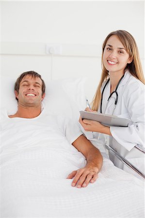 patient happy in bed - Smiling doctor standing next to a hospital bed while working with a clipboard Stock Photo - Premium Royalty-Free, Code: 6109-06007084
