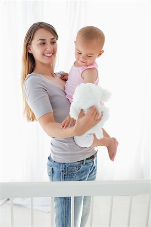 Smiling mother standing up while holding her lovely baby and a white teddy Foto de stock - Sin royalties Premium, Código: 6109-06007031