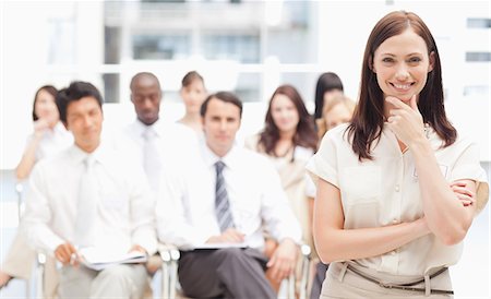 desenvolvimento profissional - Smiling brown haired businesswoman holding her hand against her chin as she stands in front of her colleagues Foto de stock - Royalty Free Premium, Número: 6109-06007270