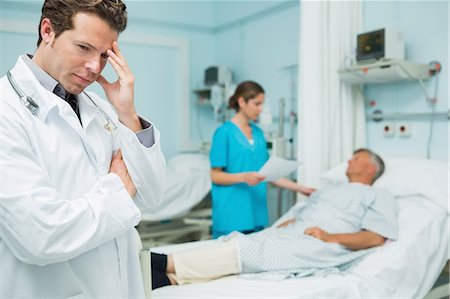 Thinking male doctor with standing in the foreground of a room Stock Photo - Premium Royalty-Free, Code: 6109-06196366