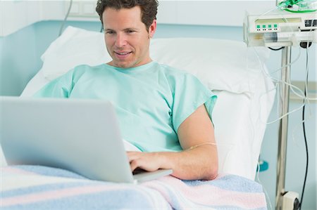 Happy male patient typing on a laptop while lying on a bed Stock Photo - Premium Royalty-Free, Code: 6109-06196311