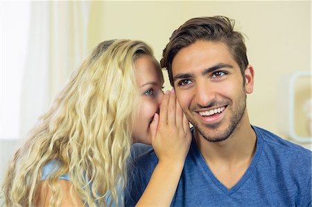 simsearch:6109-07497359,k - Blonde young woman whispering something to her boyfriend in the living room Fotografie stock - Premium Royalty-Free, Codice: 6109-07497353