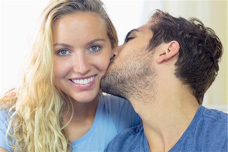 simsearch:6109-07497359,k - Blonde young woman being kissed on her cheek by her boyfriend in the living room Fotografie stock - Premium Royalty-Free, Codice: 6109-07497355