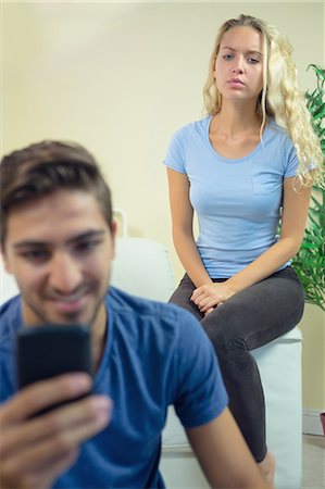 Smiling young man using his smartphone in the living room being watched by his distrustful girlffriend Foto de stock - Sin royalties Premium, Código: 6109-07497364