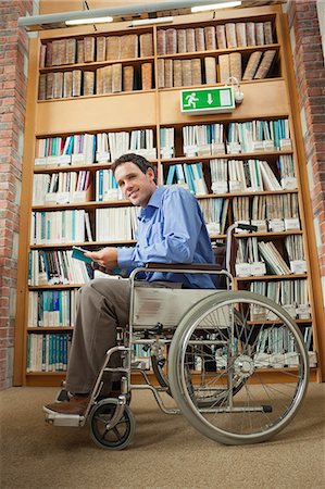 Cheerful man sitting in wheelchair holding a book in library in a college Photographie de stock - Premium Libres de Droits, Code: 6109-07497509