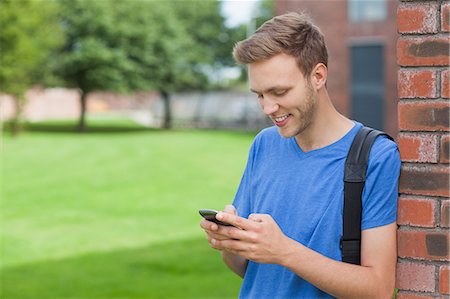 simsearch:6109-07497716,k - Smiling handsome student leaning against wall texting on college campus Foto de stock - Sin royalties Premium, Código: 6109-07497535