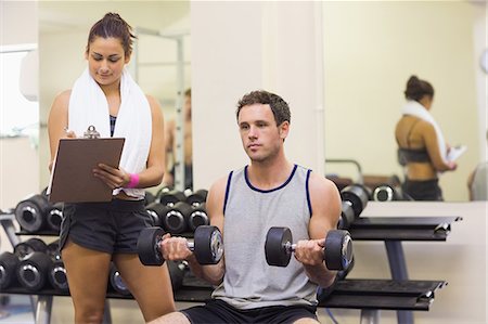 simsearch:6109-07497947,k - Trainer taking notes of attractive man lifting dumbbells in weights room of gym Foto de stock - Sin royalties Premium, Código: 6109-07497938