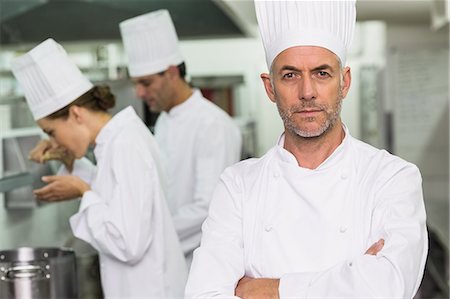 simsearch:6109-08489816,k - Serious chef looking at camera with team working behind Foto de stock - Sin royalties Premium, Código: 6109-07601048