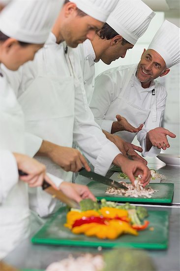 Row of trainee chefs slicing vegetables with tutor watching Stock Photo - Premium Royalty-Free, Image code: 6109-07601092