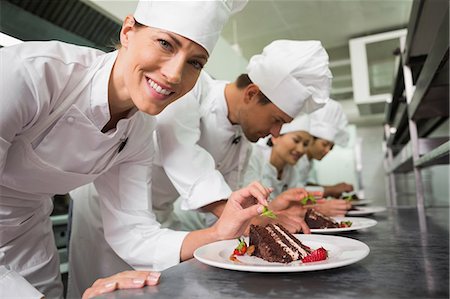 Row of chefs garnishing dessert with one smiling at camera Foto de stock - Sin royalties Premium, Código: 6109-07601101