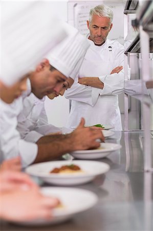 Head chef watching row of chefs garnishing spaghetti dishes with basil leaf Fotografie stock - Premium Royalty-Free, Codice: 6109-07601122