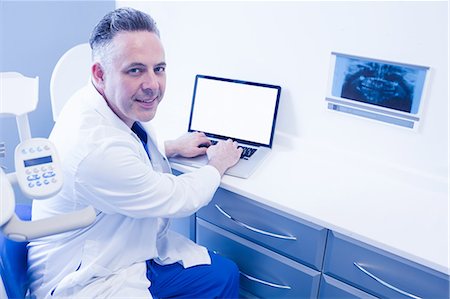 simsearch:6109-08389797,k - Smiling dentist sitting and using computer Stock Photo - Premium Royalty-Free, Code: 6109-08389783