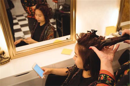 raddrizzare - Woman using mobile phone while getting her hair straightened at hair saloon Fotografie stock - Premium Royalty-Free, Codice: 6109-08764695