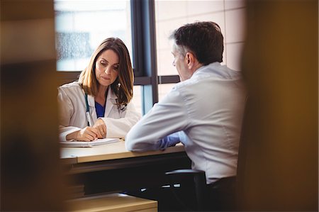 simsearch:6109-08720113,k - Female doctor sitting at her desk writing in diary in front of patient in the hospital Fotografie stock - Premium Royalty-Free, Codice: 6109-08701343