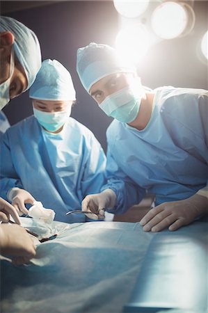 simsearch:6109-08720095,k - Surgeon looking at camera while colleagues performing operation in operation room Stock Photo - Premium Royalty-Free, Code: 6109-08720343