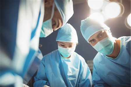 simsearch:6109-08720095,k - Surgeon looking at camera while colleagues performing operation in operation room Stock Photo - Premium Royalty-Free, Code: 6109-08720344