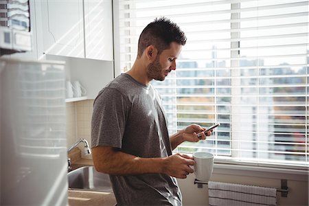 simsearch:6109-08802258,k - Man using his mobile phone while holding a coffee mug in the kitchen at home Photographie de stock - Premium Libres de Droits, Code: 6109-08802252