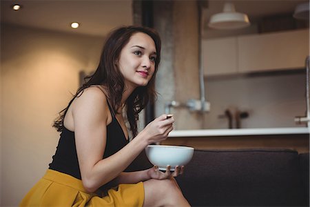 simsearch:6109-08944595,k - Woman sitting on sofa eating breakfast cereal in living room at home Photographie de stock - Premium Libres de Droits, Code: 6109-08804756