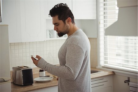 Man using mobile phone in kitchen at home Photographie de stock - Premium Libres de Droits, Code: 6109-08944666