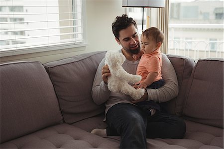 Father and baby playing with teddy bear on sofa in living room at home Stock Photo - Premium Royalty-Free, Code: 6109-08944706