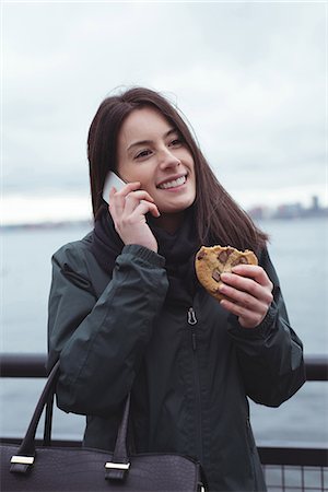 fast food city - Happy woman talking on phone while eating food against railing in city Stock Photo - Premium Royalty-Free, Code: 6109-08953639