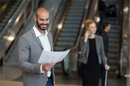 Smiling businessman standing at waiting area reading newspaper at airport terminal Stock Photo - Premium Royalty-Free, Code: 6109-08929347