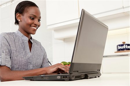 simsearch:6110-06702748,k - Young African woman working on a laptop in the kitchen, smiling Foto de stock - Royalty Free Premium, Número: 6110-06702777