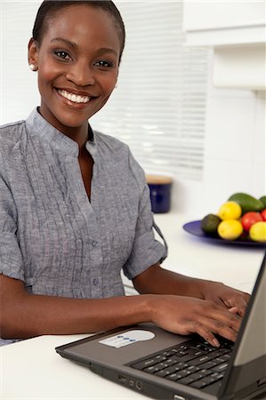 simsearch:6110-06702795,k - Young African woman working on a laptop in the kitchen, smiling at camera Foto de stock - Royalty Free Premium, Número: 6110-06702776