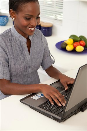 simsearch:6110-06702748,k - Young African woman working on a laptop in the kitchen, smiling Foto de stock - Royalty Free Premium, Número: 6110-06702778